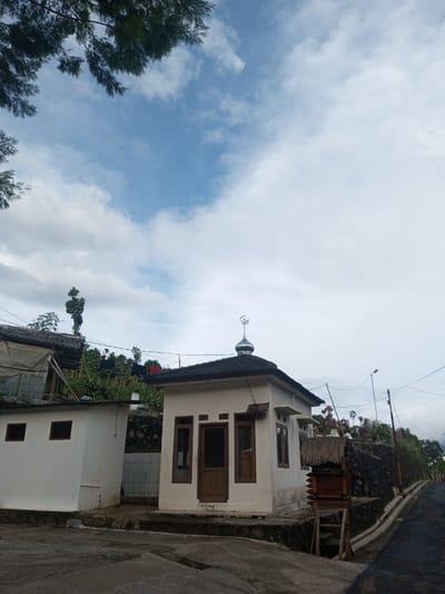 A small, white building with a silver dome and crescent moon sits near a road. Windows and a wooden door adorn the structure. To the left, another smaller, white building is attached. A stone wall with vegetation rises behind, and a utility pole stands further back. The sky is filled with white clouds, framed by green foliage at the top left. A dark road stretches into the distance on the right. A wooden structure stands adjacent to the small building.