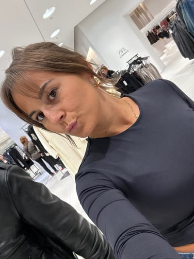 A woman with brown hair and bangs poses in a clothing store. She wears a dark long-sleeved top and gold jewelry. Racks of clothing and other shoppers are visible in the background. The store is well-lit with white walls and bright lights. A black leather jacket is partially visible in the foreground. The photo is taken at an unusual angle, with the camera positioned low and to the side of the subject.