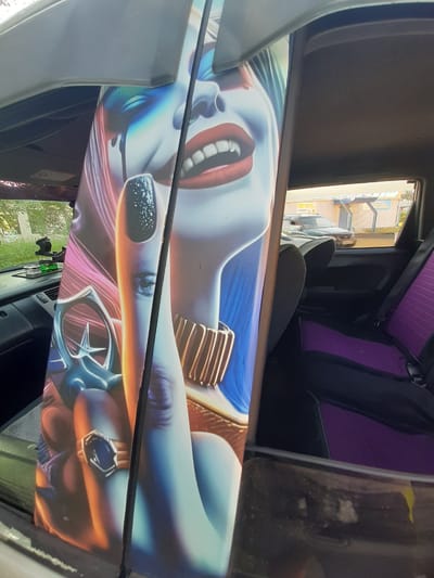 The image shows the interior of a vehicle, likely a car or van, decorated with a large airbrushed graphic. The graphic depicts a stylized, close-up image of a woman's face, reminiscent of the character Harley Quinn, with exaggerated features and vibrant colors. She has bright red lips parted to reveal white teeth, wide blue eyes, and pink and blue hair that flows around her face. She is adorned with a large golden bracelet on her arm and a prominent ring with a dark blue stone on her finger. The graphic itself is on a flat surface, possibly a door panel or dashboard, and it spans the width of the visible interior.
The vehicle's interior is partially visible, including the seats which are upholstered in a black and purple quilted fabric. The perspective suggests the photo is taken from inside the vehicle, looking towards the passenger side or rear. Outside the vehicle, through the windows, a residential area with buildings and some greenery can be seen, suggesting a daytime setting. The lighting appears to be natural daylight. No specific activity or event is taking place; the image focuses solely on the decorative artwork within the vehicle. There is no visible text in the image, other than what might be on a small sticker on the interior panel, which is not legible. The overall impression is one of personalized vehicle customization. The location context provided indicates this scene is in Shelekhov, Russia.