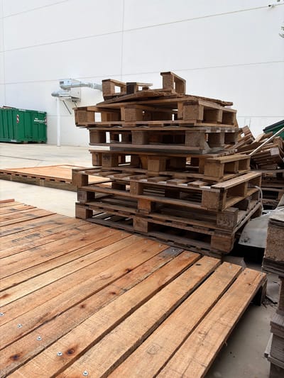 The image depicts a stack of wooden pallets and lumber, likely outdoors, in Sant Esteve Sesrovires, Spain. The pallets appear to be mostly stacked haphazardly and of varying construction. To the right, wooden boards are arranged in a row. In the background, a light-colored building wall, an electrical box, and a green container are visible. The overall environment suggests a storage area or a recycling center. There are no people visible in the image.