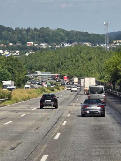 The image captures a view from behind several vehicles traveling on a multi-lane highway, presumably a Bundesautobahn, in Germany. The foreground shows the rear of a black SUV and a grey convertible. Further ahead, multiple trucks, vans, and cars are visible in various lanes, indicating moderate traffic. The highway is bordered by grassy shoulders and dense green trees on both sides. In the background, rolling hills covered with more trees form a backdrop to a residential area with scattered houses. A tall telecommunications tower is visible on a distant hill. The sky is overcast with a few scattered clouds. The scene depicts a typical highway environment on a daytime with vehicles moving along the road. The specific location is identified as Montabaur, Germany, based on the presence of a sign that appears to be a "Montabaur" exit sign, though this cannot be definitively confirmed from the image itself.