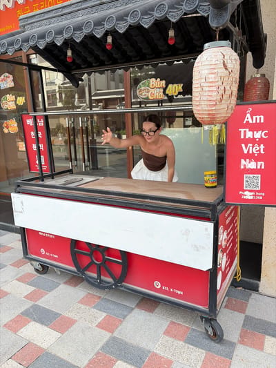 A young woman with glasses and a brown strapless top with a white skirt is standing behind a red and white food cart. She is reaching out her right hand toward the viewer. The food cart has a black metal frame and a wooden countertop. There are various signs on and around the cart, including one with Vietnamese text and a QR code, and another with images of food. A large, slightly damaged paper lantern hangs from the top of the cart. The cart is positioned on a checkered tile surface. In the background, there are reflections of buildings in a glass window.