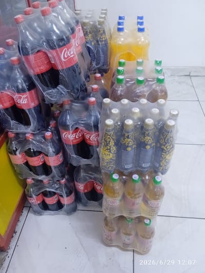 The image shows an indoor scene, likely a store or storage area, with several multi-packs of bottled beverages. In the foreground and to the left, there are multiple packs of Coca-Cola bottles. The bottles are dark brown, and the distinctive red Coca-Cola branding is visible. To the right and towards the back, there are packs of bottles containing yellow and amber-colored liquids, some with blue caps and others with green caps. One set of these bottles has ornate black and gold labels. The floor is tiled in a light color, and there is a yellow wall to the far left. The lighting is neutral. There are no people or animals visible in the image. The time stamp "2026/6/29 12:07" is visible in the lower right corner. This scene is not identifiable as being in the city of Ak-Suu, Kyrgyzstan, or any specific geographic location.