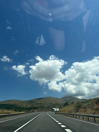 A road view from a vehicle, looking ahead on a highway. The highway has two lanes in the direction of travel, separated by white dashed lines. A metal guardrail runs along the right side of the road. In the distance, on the right side of the highway, there are trucks. The landscape on either side of the highway consists of dry, brown hills with sparse vegetation and some trees. In the background, there are mountains under a clear blue sky with scattered white clouds. The sky shows reflections of the interior of the vehicle from which the photo was taken. The location is identified as the city of El Espinar, Spain.