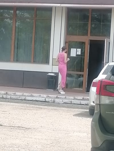 A woman in a pink dress is standing near the entrance of a building. She is facing away from the camera, and appears to be holding a phone. The building has large windows and a glass door. A dark green car is parked to the right of the entrance. The ground in front of the building is paved with gravel. The image is blurry and appears to have been taken with a low-resolution camera. The location is the city of Lesozavodsk, Russia.