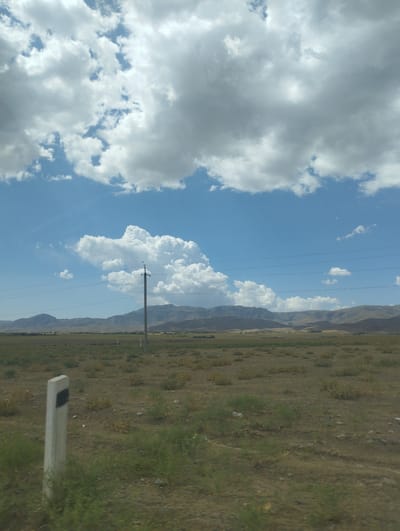 The image shows a wide, open landscape under a partly cloudy sky. In the foreground, a light-colored marker post with a black band is visible on the left. The ground is covered in dry grass and scattered bushes, with some areas appearing bare and dusty. In the mid-ground, a power pole with wires stretches across the frame, and further in the distance, a range of hills or mountains forms the horizon. The sky is a vibrant blue with large, puffy white clouds, some of which are tinged with gray, suggesting recent or impending rain. The overall impression is one of a rural, undeveloped area. This scene is consistent with the natural landscape found in Forish QFY, Uzbekistan, which is known for its steppe and mountainous terrain. There are no people or specific buildings clearly identifiable in the image, adding to the sense of a vast and natural environment.