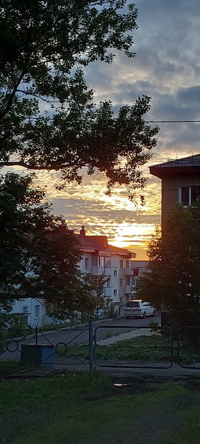 The image depicts a sunset scene in Aleksandrovsk-Sakhalinskiy, Russia. In the foreground, there is a dark grassy area with a metal fence. Behind the fence, a road curves, bordered by green grass and trees. Several multi-story residential buildings with white facades and red roofs are visible in the middle ground. A white van is parked on the road. The sky is filled with dramatic clouds illuminated by the setting sun, which casts a warm golden glow. The overall impression is that of a residential neighborhood at dusk.