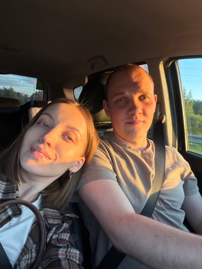 The image depicts two people in a car, likely on a road trip, given the presence of seatbelts and the view from the windows. One person, a man, is wearing a light brown t-shirt and has a bald head. The other person, a woman, is wearing a plaid shirt over a white t-shirt and has shoulder-length brown hair and earrings. They are situated inside the car with the camera pointed upwards at them. The presence of the sunroof suggests a sunny or bright day. The location is unlikely to be Rogachovo, Russia, due to a lack of environmental context.