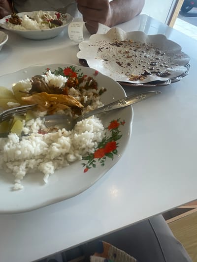 In a brightly lit indoor setting, likely a restaurant in Medan, Indonesia, a partially consumed meal is presented on a white table. The focal point is a white ceramic plate with a floral pattern, featuring a generous portion of white rice, a piece of cooked chicken, and what appears to be some form of vegetable and sauce. A spoon and fork rest on the plate, indicating recent activity. To the right, a brown woven basket holds a fluted paper liner, stained with remnants of sauce and food, suggesting it may have held another dish or served as a disposable serving surface. In the background, another white bowl filled with rice and toppings is visible, and the edge of a receipt is also present. The scene suggests a casual dining experience, possibly after a meal has been enjoyed or is in progress.