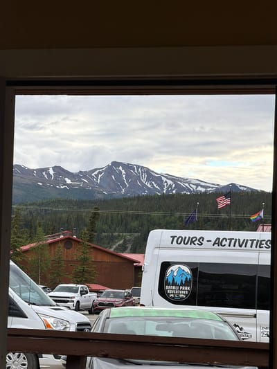 The image shows a view of Denali National Park and Preserve in Healy, Alaska, United States, from a window or opening. In the foreground, there are several vehicles, including a white van with "TOURS - ACTIVITIES" and "DENALI PARK ADVENTURES ALASKA" branding, and a white pickup truck with "CHEVROLET" visible on the grille. A red SUV is also present. Beyond the vehicles, a wooden building with a red roof is visible, surrounded by evergreen trees. The background features a majestic mountain range with snow-capped peaks and a forest of trees. Several flags are flying, including the American flag, the Alaskan flag, and a rainbow flag. The sky is overcast with clouds.
