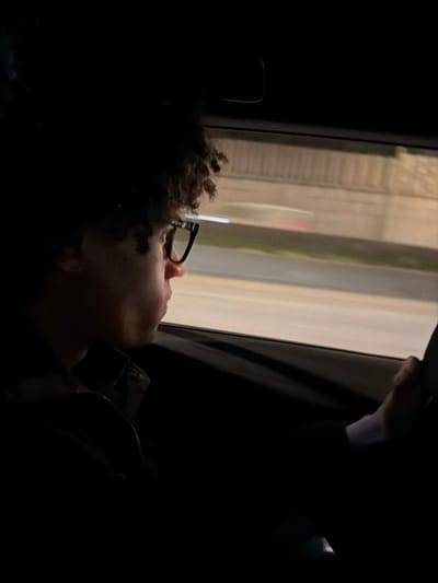 A person with curly hair and glasses gazes out a window in a dark vehicle. The window reflects a blurred urban scene, possibly buildings or structures. A hand is visible near the top of the window frame. The person is wearing a jacket, with details like a zipper and collar discernible. The overall image has a somber, contemplative mood due to the low lighting.