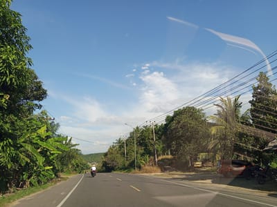The image shows a road in Blangkarieng, Indonesia, during daylight. A person on a motorcycle is traveling away from the viewer on the asphalt road, which has a yellow dividing line. The road is lined with dense vegetation on both sides, including banana trees and other foliage. Utility poles and wires run along the right side of the road. A vehicle and motorcycles are parked on the right shoulder. The sky is blue with scattered clouds. There is a reflection on the camera lens.