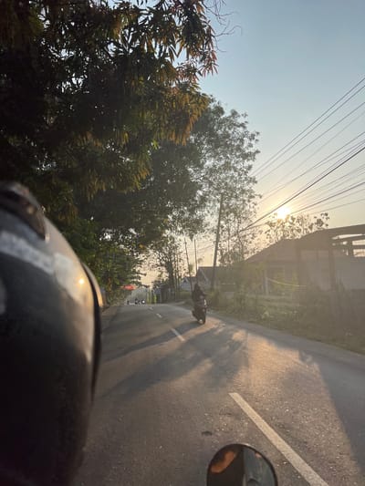 The image depicts a scenic road in Klari, Indonesia, bathed in the warm glow of a setting sun. The perspective is from the viewpoint of someone riding a motorcycle, with the helmet partially visible in the foreground. The road stretches forward, lined with lush green trees and vegetation on either side. A few motorcycles are seen in the distance, creating a sense of motion. Power lines crisscross the sky, adding to the rural atmosphere. The sunlight casts long shadows on the road, emphasizing the late hour. The overall impression is one of a peaceful and picturesque journey through the Indonesian countryside.