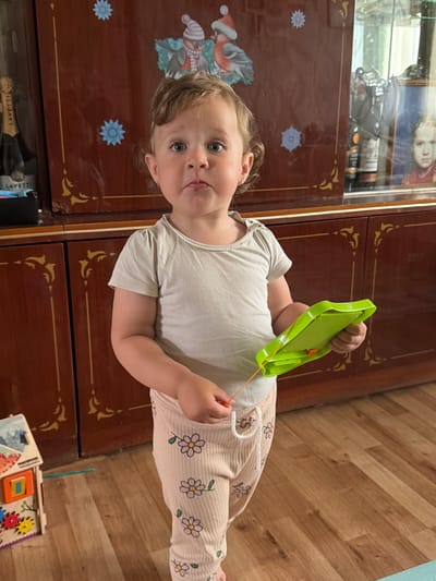 A young child with curly brown hair and blue eyes stands in front of a wooden cabinet. The child is wearing a light gray t-shirt and beige pants with a floral pattern. The child is holding a bright green toy with an orange string. The cabinet is dark brown with decorative gold accents. On the cabinet, there are two light blue snowflake decorations and a picture of birds. To the left of the child, a bottle of champagne is visible on a shelf. To the right, the cabinet has a glass door, revealing a shelf with bottles and a framed picture of a young girl. The floor is made of wooden planks. The scene appears to be an indoor residential setting, possibly in Komsomolskiy, Russia, given the context.