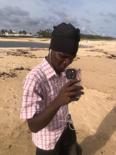 The image shows a person, presumably male, taking a selfie with a smartphone on a beach in Akodo, Nigeria. The person is wearing a black cap and a red-and-white checkered shirt and has earphones plugged in. The beach is sandy, with some seaweed or debris scattered around. In the background, there is a line of palm trees and buildings, and the sky is overcast. Two individuals can be seen on the horizon. The person appears to be standing still while taking the photo.
