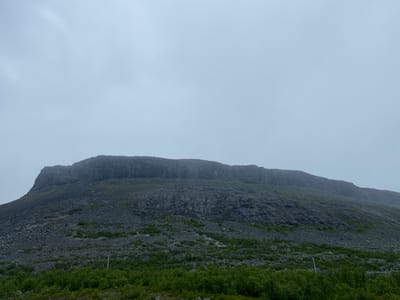 The image depicts a vast, rocky mountain under a cloudy sky. The mountain's summit is flat and layered, resembling a mesa. Its slopes are covered in scree and sparse green vegetation, primarily low-lying bushes. A single white pole, possibly a marker or part of a fence, is visible on the lower slope amidst the greenery. The overall atmosphere is somber and natural, suggesting a remote, mountainous region. The provided context indicates the location is Kiruna, Sweden.