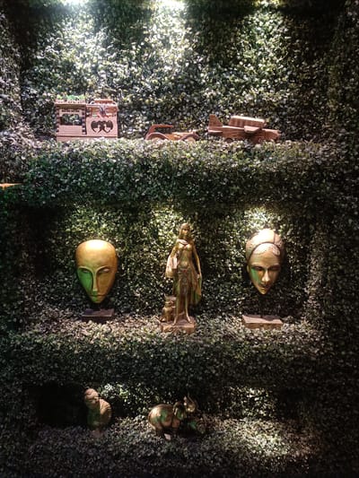 The image shows a display of decorative objects against a backdrop of dense green foliage, likely artificial. The display is organized on shelves, with spotlights illuminating the items.
On the top shelf, there are three wooden objects. The leftmost object appears to be a miniature building with decorative carvings and a small plant on top. To its right is a wooden representation of a chariot or cart with wheels. Further to the right is a wooden model of an airplane with visible wings and a cockpit.
The middle shelf features three sculptures. On the left is a large, golden mask with an elongated face and stylized features. In the center stands a golden statue of a female figure, possibly a warrior or goddess, adorned with intricate clothing and jewelry, holding a smaller figure of an animal at her feet. To the right of this statue is another golden mask, this one with a more ornate headdress and facial details.
The bottom shelf displays two smaller golden sculptures. On the left is a seated figure, possibly a monkey or a child. To its right is a sculpture of an elephant with its trunk raised.
The overall scene is dimly lit, with the spotlights creating pools of light and shadow on the foliage and the displayed objects, suggesting an indoor display, possibly in a shop or a curated exhibition. The time of day cannot be determined from the image. No text is visible. The setting appears to be a decorative display of art pieces, possibly inspired by various cultural motifs, presented in a visually appealing manner. The location context of Kurnool, India, might suggest a local craft or artistic representation.