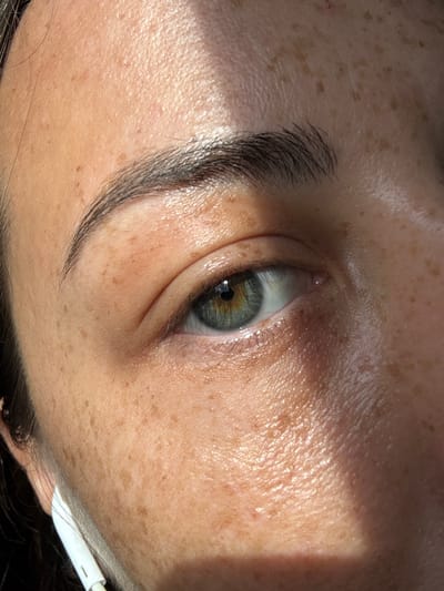 A close-up, eye-level shot focuses on a portion of a person's face, primarily the right eye and surrounding skin. The individual has fair skin with numerous freckles scattered across the forehead, cheek, and under the eye. A prominent eyebrow, dark and well-defined, arches above the eye. The eye itself is a striking shade of green with flecks of gold and blue, and the pupil is a deep black. Eyelashes are visible, framing the eye.
The skin appears slightly dewy and reflects the sunlight. A diagonal shadow, cast by an unseen object, cuts across the forehead and eyelid, creating a contrast between illuminated and shadowed areas. In the lower left corner of the frame, a white earbud with a cord is partially visible, suggesting the person is wearing earphones. The background is out of focus, but hints of dark hair are visible along the left edge of the image. The overall impression is one of natural light and close-up detail, emphasizing the texture and features of the skin and eye.
The location in the city of Phan Son, Vietnam is not discernible from the provided image.