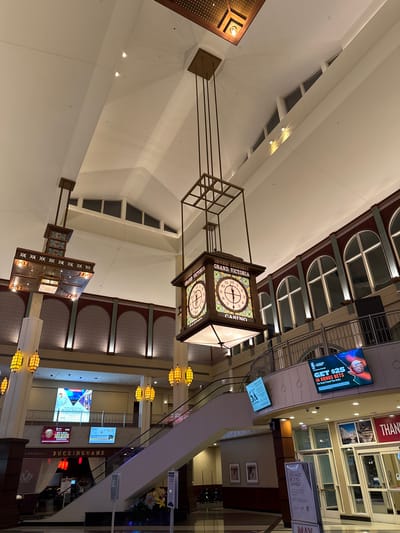 This indoor shot showcases a multi-level building, likely a casino or a large entertainment complex, located in Elgin, United States of America. The image is taken from a lower vantage point, looking upwards. The central focal point is a large, decorative clock hanging from the ceiling. The clock has a square frame and features a clock face on each side, displaying "Grand Victoria Casino" around the clock.
The building features a mix of architectural styles, with arched windows on the upper levels and a modern, open design. There are displays and advertisements for the casino, including a promotional sign offering "$25 in Bonus Bets." An escalator runs diagonally across the frame, and a restaurant named "Buckinghams Steakhouse" is visible.
The environment is well-lit, with a mix of natural and artificial lighting. There are no people visible in the immediate foreground, suggesting the image was taken when the area was not heavily populated.