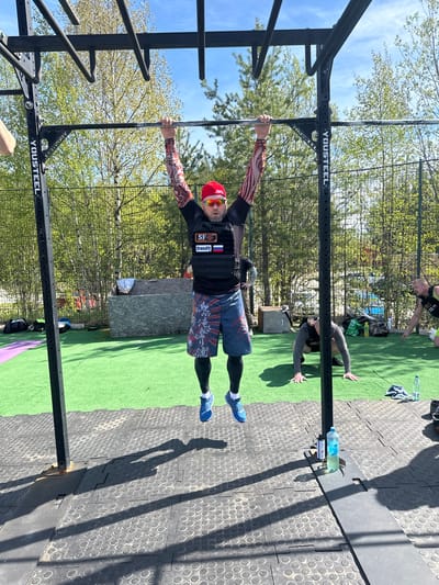 The image shows a man performing a horizontal handstand on a pull-up bar in an outdoor fitness area. The man is wearing a red cap, sunglasses, a black shirt with CrossFit patches, and patterned sleeves and shorts. He is positioned with his body parallel to the ground and arms fully extended, gripping the bar. The pull-up bar is part of a black metal workout structure labeled "YOUSTEEL".

The setting includes an outdoor workout area with a combination of artificial green turf and black rubber mats. In the background, there are trees suggesting a park or wooded area. Other individuals are present in the background, appearing to be engaged in physical activity. A water bottle is visible near one of the metal supports. The scene is well-lit, indicating a sunny day. The flag of Russia can be seen on the person's chest.
There is a green turf and a black rubber mat in the area. Based on the text on the apparel, the image is likely in Polarnie Zori, Russia.