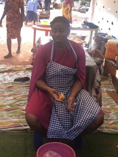 A woman is seated at a table in what appears to be an outdoor market setting. The woman wears a dark pink dress with a blue and white checkered apron. She is holding what looks like a piece of bread in her hands. She is sitting on a blue stool. A pink plastic basin is on the ground next to her. The table is covered with striped fabric. The foreground shows a sandy surface with two partially visible people. A woman is standing beside an orange plastic table with several blue skirted people behind it. Based on visual cues, the location is Nteje, Nigeria.