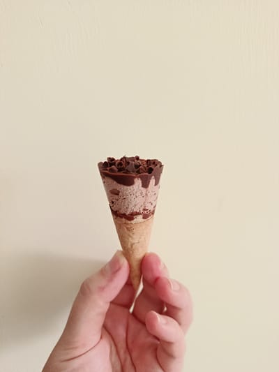 A right hand holds a small, chocolate-flavored ice cream cone upright against a plain, off-white background. The ice cream appears to be a mottled light brown and pinkish hue, possibly a mix of chocolate and another flavor, with a visible texture suggesting a frozen dessert. The top rim of the ice cream is covered in a dark brown, solidified chocolate coating, above which small, curled chocolate shavings or flakes are scattered. The cone itself is a light brown, waffle-textured sugar cone. The hand, with light skin tone, grasps the lower portion of the cone, with the thumb and index finger prominently visible. Fingernails are short and unpainted. The background is a uniform, solid cream or very light beige color, displaying minor surface irregularities consistent with a painted wall. The scene is depicted indoors or in an area with a simple backdrop, reportedly located in the city of Águilas, Spain.