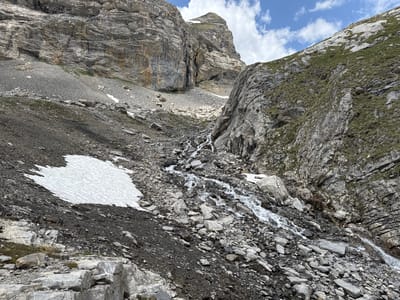 The image depicts a rugged mountain landscape, characteristic of the Swiss Alps, with a rocky incline and a small stream of water flowing downwards. Patches of snow are visible on the slopes, suggesting a high altitude or a recent snowfall. The terrain is predominantly rocky, with scattered vegetation, primarily grass, clinging to the surfaces. Massive rock formations dominate the upper parts of the scene, with layers of stone indicating geological history. The sky above is partly cloudy, with blue sky visible between fluffy white clouds. The overall impression is one of a wild and natural environment, untouched by significant human intervention. Given the context provided, this scene is likely located in the vicinity of Leukerbad, Switzerland, a region known for its mountainous terrain and thermal baths.