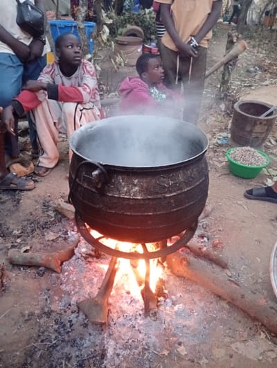 A large, black cauldron steams over an open fire supported by a metal stand and wooden logs. Surrounding the fire are several people, including two young children. One child in a pink hooded sweatshirt is partially obscured by steam. A green bowl of beans sits beside a wooden bucket. Sandals are visible on the feet of those nearby, set on a ground scattered with ash and dry leaves.