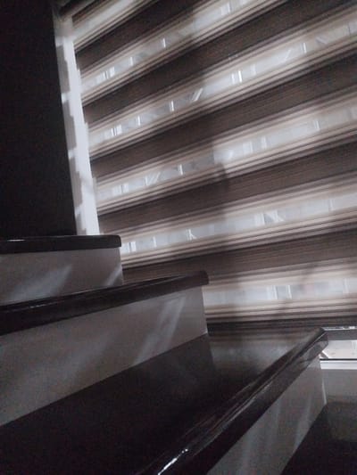 A partially visible indoor staircase is shown in the foreground, with the steps covered in a dark, possibly varnished wood. The staircase railing is also dark. In the background, a window is covered by horizontal blinds with alternating opaque and sheer stripes, creating a patterned effect. The blinds are in shades of brown and beige. The overall scene appears to be an interior shot, possibly in a residential building. The location is described as Социализм, Kyrgyzstan.