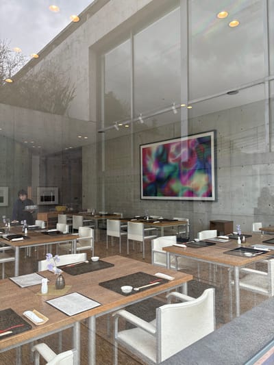 The image depicts a view from inside a glass-walled building. The building's interior is modern and minimal, with a concrete ceiling and exposed beams. The ceiling is lit by a series of recessed lights. A large abstract painting hangs on the wall, featuring a multi-colored swirling pattern. Several tables are set up for dining, with white chairs and linens. A woman in a white shirt and black pants stands near one of the tables, arranging flowers. The scene is located on the island of Naoshima, Japan, at the coordinates 34.445 latitude and 133.991 longitude. The image is shot from a low angle, looking up towards the ceiling. The glass wall acts as a mirror, reflecting the surrounding environment. The reflection shows trees and the sky.