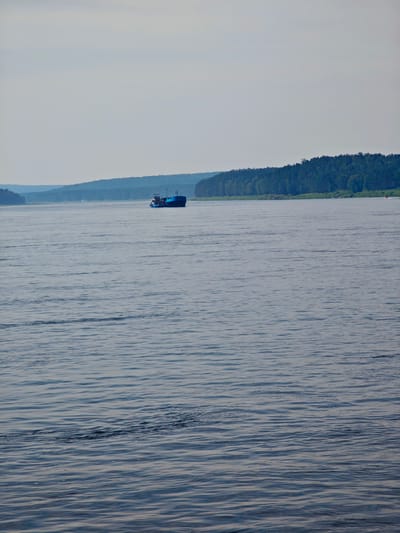 The image depicts a wide, calm river with gentle ripples reflecting the overcast sky. In the middle distance, a blue barge with several structures on its deck is moving across the water. The riverbanks are lined with dense green trees, forming a natural, forested landscape. In the far distance, layered hills fade into a hazy sky, suggesting a broad, open expanse. The scene is serene and natural, with no prominent urban structures visible. The location is identified as the city of Podporozh'e, Russia, which is situated on the Svir River, known for its navigable waterways and surrounding forests.