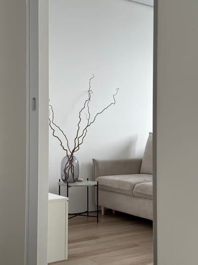 The scene depicts an interior view within a structure located in Башкултаево, Russia. The image captures a section of a room through an architectural opening, bordered by light-colored vertical wall or door frame elements on both the left and right sides. The primary visible environment features plain white walls and a light-colored wooden floor. A large, grey-tinted glass vase, containing multiple bare, gnarled tree branches, is centrally positioned on a small, round side table. This table has a light grey top and black metal legs. A small, smooth, light-colored stone is also visible on the table adjacent to the vase. To the right, a portion of a light beige fabric sofa is visible, equipped with light-colored wooden feet. No individuals are present, and no actions or interactions are depicted. The lighting is uniform, suggesting an interior illumination or diffused natural light source outside the frame.