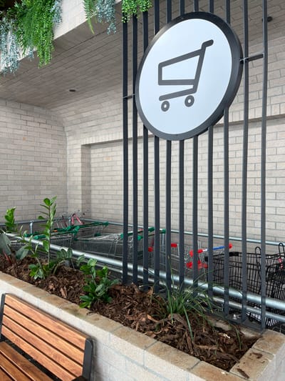 This image depicts a brightly lit outdoor area, likely a trolley bay or collection point associated with a retail establishment in Indooroopilly, Australia.

The central focus is a circular white sign with a thick black rim, prominently displaying a minimalist black outline icon of a shopping cart. The sign hangs vertically, attached to one of the horizontal bars of a black metal slatted fence. This fence runs horizontally across the middle of the frame, serving as a barrier.

Behind the fence, numerous metal shopping trolleys are stacked and stored, some identifiable by their red or green handles. To the right of the fence, there is a raised planter box filled with dark mulch, nurturing various green plants, including a tall, spiky, grass-like plant and a leafy plant with wider foliage. A section of a modern wooden slatted bench with a black metal frame is partially visible on the far right, adjacent to the planter.

The background consists of light-colored brick walls, featuring different patterns and sections, suggesting commercial architecture. The ground is paved with light-toned bricks or tiles. On the far left, lush green plants are visible, adding to the outdoor aesthetic. The scene is devoid of people, conveying a sense of quiet functionality. The clear, even lighting indicates it is daytime. No discernible text is present, only the graphical representation of a shopping cart.