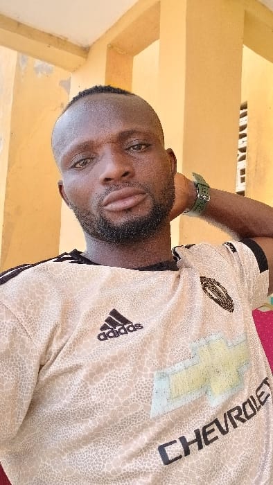 The image displays a close-up portrait of an adult male located in Bashar, Nigeria. The male, who has dark skin, short dark hair, a full beard, and a mustache, is looking directly at the camera. He is wearing a light beige short-sleeved t-shirt with a textured pattern, a black Adidas logo on the upper left chest, and a faded yellow Chevrolet bow-tie emblem with the word "CHEVROLET" printed in dark letters across the front. A dark green wristband is visible on his left wrist, and his left arm is bent, with his hand positioned behind his head. The background consists of a building structure painted in a faded yellow or light orange color, featuring vertical pillars and a section of white louvers or slats on the right. The wall on the left shows minor paint wear.