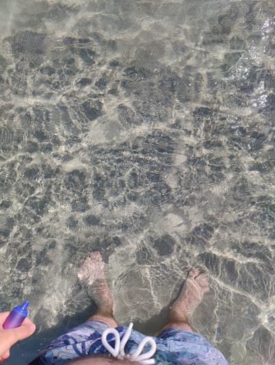 This is an outdoor, top-down, medium shot image of a person's legs and feet submerged in shallow, clear ocean water at a beach. The person is wearing blue patterned swim trunks. The bottom of the ocean is visible through the water, appearing to be composed of sand and small stones, with sunlight reflecting off the water's surface, creating patterns of light and shadow. The person is holding a purple vape pen in their left hand, which is partially visible at the bottom left of the frame. The scene is likely in the city of Aegina, Greece, given the context.