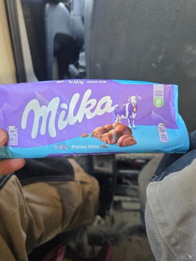 A human hand is holding a Milka Bubbly Alpine Milk chocolate bar in the interior of a vehicle within the city of Opalchenets, Bulgaria. The chocolate bar, encased in its distinctive purple and light blue wrapper, features the white "Milka" logo, an Alpine cow illustration, and depictions of aerated chocolate pieces. Text visible on the wrapper includes "bubbly," "Alpine Milk," "100% Alpine Milk," and an emblem for "SUSTAINABLY SOURCED COCOA." The hand, partially visible on the left, holds the bar horizontally across the frame. Below the chocolate bar, the person's legs are visible, clothed in brown/khaki fabric on the left and a light blue and white striped fabric, possibly denim, on the right. The immediate background behind the chocolate bar consists of dark gray or black textured upholstery, consistent with a car seat. Further back, the blurry interior of a vehicle is discernible, suggesting a confined space.