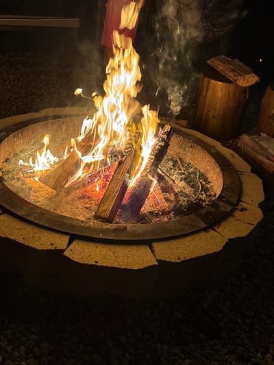A fire burns brightly in a circular stone fire pit at night, casting a warm glow on the surrounding gravel and logs. Several pieces of firewood are arranged in the pit, with flames leaping upwards and wisps of smoke drifting into the darkness. In the background, a person in a pink shirt stands partially obscured, and two large logs are stacked next to the fire pit. The scene is set in the Town of Dell Prairie, United States of America.