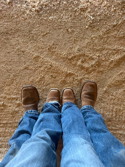 An overhead, downward-facing perspective captures the lower extremities of two individuals, an adult and a child, standing on a ground surface composed of light brown, granular material consistent with wood shavings or sawdust. Both individuals wear blue denim jeans and square-toed, brown leather cowboy boots. The adult's larger, darker brown boots are positioned behind the child's smaller, lighter brown boots. The child's blue jeans exhibit frayed cuffs. The scene suggests an equestrian or stable environment within Rancho Murieta, United States.