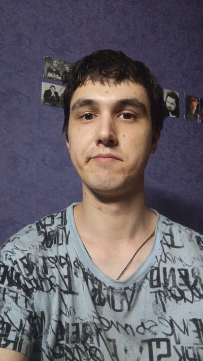 A young man with dark, tousled hair and fair skin is looking directly at the camera. He has brown eyes, a slightly pursed mouth, and a few small blemishes on his face and neck. He is wearing a light blue t-shirt with a black graffiti-like pattern. A thin, dark cord necklace is visible around his neck. The background is a textured purple wall adorned with several small black and white photographs. The overall impression is of a casual, indoor portrait. The image is a selfie, taken from a slightly low angle. There is no indication of the specific location within Gryazi, Russia.