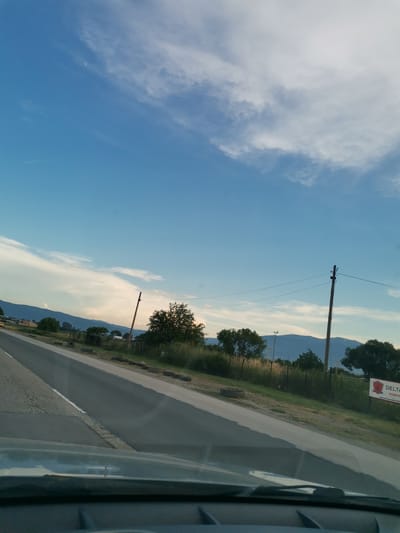 The image shows a clear blue sky with some wispy clouds above a rural landscape. In the foreground, a portion of a car's windshield and dashboard is visible, suggesting the photo was taken from inside a vehicle. A road runs diagonally through the scene, bordered by a grassy verge and some vegetation. In the middle ground, a utility pole with wires stretches across the frame, and several trees are scattered around. Beyond the trees, a range of mountains forms the horizon. In the far right, a red and white sign with text in Cyrillic script is partially visible. The overall scene appears to be a roadside view on a sunny day. There are no people or animals clearly visible in the image.