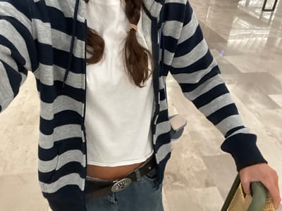 The scene is set in an indoor environment in San Miguel Jaltocan, Mexico, featuring light-colored, reflective flooring. The image captures a close-up, downward-angled view of a single individual, partially obscuring the head and legs. The subject wears a white crop top, a grey and navy blue horizontal-striped zip-up hoodie, and blue jeans. A dark brown belt with a circular, silver-toned buckle is visible at the waistline. Long, brown hair is styled into at least one braid, secured with a light-colored elastic band near the right shoulder. A small, light-colored object, possibly a shoe, is partially contained within the left pocket of the hoodie. The individual's right hand grips a green, textured handle, consistent with that of a luggage item or cart. The action depicted is the individual holding and presumably moving this object. No other individuals are present in the visible frame.