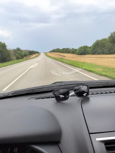 The image captures a view from inside a vehicle looking out through the windshield. In the foreground, resting on the dashboard, is a pair of black sunglasses. The windshield wipers are visible. The road ahead stretches into the distance, curving slightly and bordered by fields of dry, golden grass and green trees. The sky is overcast with a pale, cloudy sky. The location is identified as Kruzhilinskii, Russia.