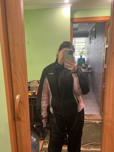 A woman is taking a mirror selfie, capturing herself from the chest up. She is wearing a black and pink jacket with black pants. The reflection also shows a glimpse of the room, which includes a green wall, a hallway, a patterned rug, and various objects such as boxes and shoes. The image was taken indoors in the city of Cherliany, Ukraine.