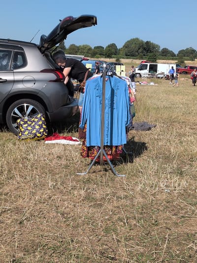 A person is loading items into the trunk of a gray car at an outdoor market. The car's trunk is open, and the person is leaning into it. A rack of clothing hangs in the foreground, and other people and vehicles are visible in the background, suggesting a car boot sale. The ground is covered with dry grass, and the sky is clear and blue. The location is the village of Claverley, United Kingdom.