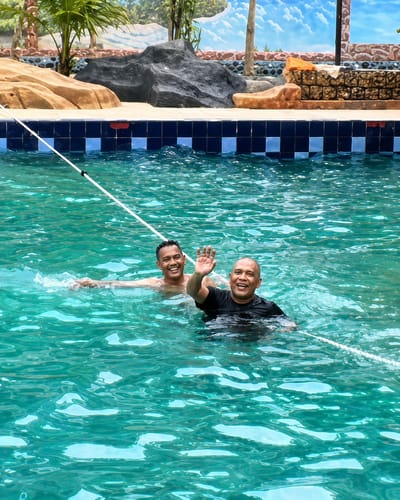 Two adult males are in a swimming pool, one in the foreground and one behind him. The man in the foreground is wearing a black t-shirt and is smiling and waving at the camera. The man behind him is wearing a white shirt and is also smiling. The water in the pool is a clear turquoise color. The pool is outdoors and surrounded by a tiled edge. In the background, there is a mural of a landscape with mountains and a blue sky. There are also some decorative rocks and palm trees around the pool. The image appears to have been taken on a sunny day. The location of Eampat Kwalamencirim, Indonesia, is not visually identifiable from the image itself.