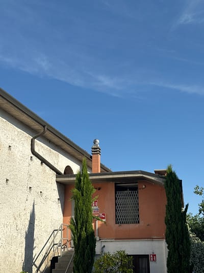 The image depicts a building in Villafranca di Verona, Italy. The building has a light-colored, textured exterior wall. A gutter runs along the roofline. An orange section of the building is visible with a window protected by a metal grate. Green foliage, likely trees or bushes, are present near the top of the building. The sky is bright blue with some wispy clouds. A chimney is also visible. There are some fire safety signs on the wall. Metal stairs are visible on the right.