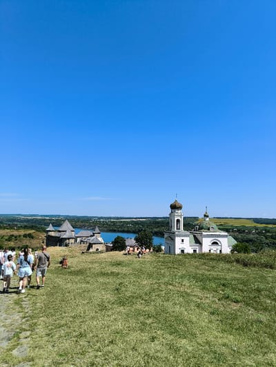 The image depicts an outdoor, daytime scene in Khotyn, Ukraine, featuring historical structures and a natural landscape under a clear blue sky.

In the foreground, a grassy hillside slopes downwards from the viewer. A cobblestone path is visible on the left, occupied by a group of approximately five individuals walking away from the camera, heading downhill. These individuals are dressed in casual summer attire, including shorts and t-shirts.

The midground is dominated by two primary architectural subjects. To the right, a white-painted church building with a prominent bell tower rises. The church features a green-domed main roof and the bell tower is topped with a smaller, lighter-colored dome and a cross. Multiple arched openings are visible on the bell tower. To the left of the church, and further downhill towards a large body of water, stands the Khotyn Fortress. This extensive stone structure comprises multiple towers with pointed or conical roofs and interconnected walls. The large body of water, identifiable as the Dniester River, stretches across the midground behind the fortress and partially behind the church. Scattered smaller buildings and trees are visible between the church and fortress, and along the riverbank. Several small figures are discernible walking near the fortress and river.

The background consists of a vast, unblemished clear blue sky occupying the upper third of the image. Beyond the Dniester River, the landscape transitions into distant rolling hills, characterized by patches of green foliage and yellow fields, extending to the horizon.