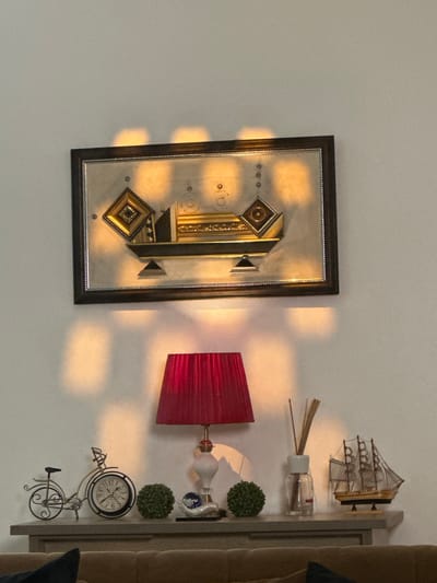 The image depicts an indoor scene, likely a living room, with a wall adorned by a rectangular framed artwork. The artwork features a geometric design with metallic accents in gold and silver, reflecting the warm light of what appears to be sunset or artificial lighting. Below the artwork, on a narrow shelf or mantle, several decorative objects are arranged.
From left to right, these objects include a miniature metal bicycle, a clock designed to resemble a bicycle wheel, a small green spherical plant, a white and red table lamp with a pleated shade, a diffuser with reeds, and a model of a wooden sailing ship. The shelf is positioned above a piece of brown furniture, possibly a sofa.
The lighting creates a dramatic effect, with distinct rectangular shadows cast on the wall above and to the left of the shelf, originating from an unseen light source or window. The overall ambiance is one of a decorated interior space with a focus on decorative items and the play of light and shadow.
While the image is detailed, it does not contain any visual information that allows for the specific identification of its location within Pakuro, Nigeria. The scene is generic to many interior decorations and lacks any identifying geographical markers or cultural specificities of the region.