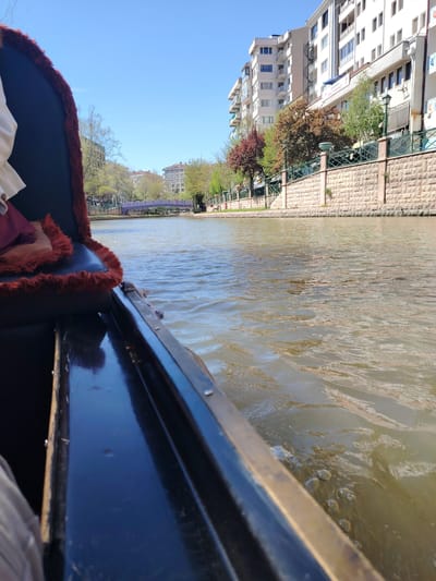 This media file presents a first-person perspective from a boat, resembling a gondola, navigating a canal or river in an urban setting. The boat's interior, visible on the left, features dark blue material with a plush, red fur-like trim. A person's arm and a glimpse of clothing are visible within the boat.
The waterway itself appears greenish-brown with ripples, indicating gentle movement. Along the right bank, a decorative stone-faced wall with a dark metal fence and streetlights separates the water from the land. Behind this wall, trees with a mix of green, red, and brown leaves suggest either spring or early autumn.
Several multi-story residential or commercial buildings, predominantly light-colored, line the bank. On one of these buildings, a white sign with blue text is visible, reading "ESKİŞEHİR GENÇLİK MERKEZİ" (Eskişehir Youth Center) followed by "MA". In the distance, a distinctive purple or lilac-colored arched bridge crosses the canal, with more urban infrastructure beyond.
The sky is clear and bright blue, indicating a sunny day. The scene depicts a calm, recreational activity in an urbanized landscape, specifically in Eskişehir, Turkey, as confirmed by the visible signage.