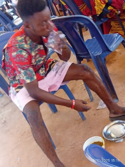 A man in a colorful shirt and pink shorts sits on a blue plastic chair, drinking from a plastic bag. He is wearing a red bracelet and sandals. Nearby, on the ground, are two metal dishes. One contains a white substance, and the other holds a brown sauce. The ground appears to be dirt or sand. Other blue plastic chairs are visible in the background. The date "19/12/2025 15:16" is stamped at the bottom right corner of the image.